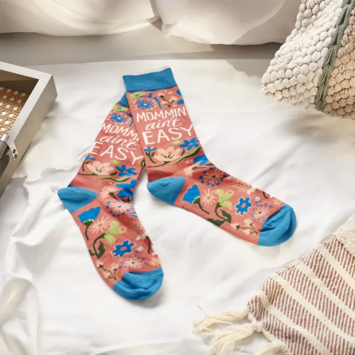 Mother's Day Collection A pair of colorful woven socks from our Mother's Day Collection displaying unique hand illustrated designs of flowers with a fun mom-themed sentiment that reads "Mommin' Ain't Easy." Socks feature a one size fits most design made with cotton, nylon, and spandex. Our Mommin' Ain't Easy socks come with a header card and a clear plastic hanger for display. A fun Mother's Day gift and accessory perfect for everyday self-expression.